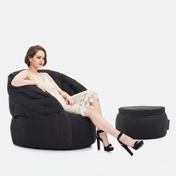Valkoiseen pitsimekkoon ja mustiin korkokenkiin pukeutunut nainen istuu suurella, tummalla Memory Foam Butterfly -sohvalla jalat ristissä. Hänen viereensä on asetettu sopiva pyöreä ottomaani, ja kaikki tämä on valkoista taustaa vasten.
