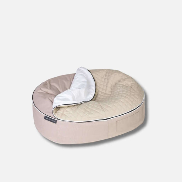 Vaaleanharmaalla pohjalla beige, ovaalinmuotoinen allergivennlig hundeseng, jonka toisella puolella on quiltet topptrekk ja valkoinen, osittain taitettu peitto.