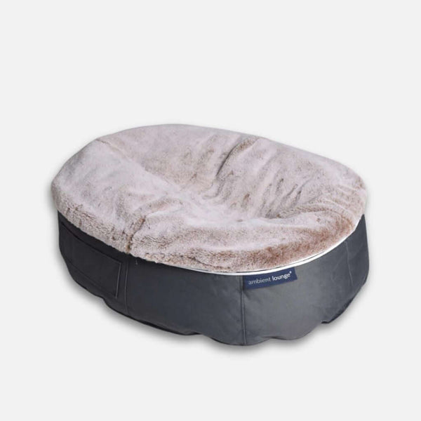 Pehmeä, ovaalin muotoinen hundeseng, jossa on muhkea, vaaleanbeige tyyny ja tummanharmaa kangasalusta, ja jonka sivussa on pieni Ambient Lounge -merkki, joka takaa täydellisen mukavuuden hundille.