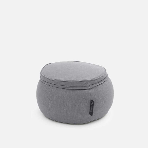 Pyöreä, vaaleanharmaa kankainen puffi, jossa on pehmeä, muistivaahtomuovipehmustettu päällinen ja pieni musta lappu sivussa, valkoista taustaa vasten.