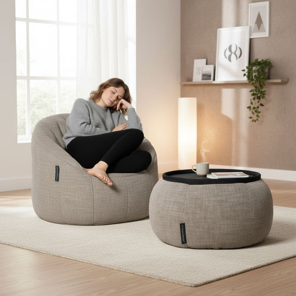 Harmaaseen villapaitaan pukeutunut nainen istuu rennosti Ambient Lounge Cove Package -papusäkkituolissa silmät kiinni. Sopiva ottomaani, jossa on tarjotin ja kuppi, täydentää viihtyisän, modernin olohuoneen mukavan møblerin.