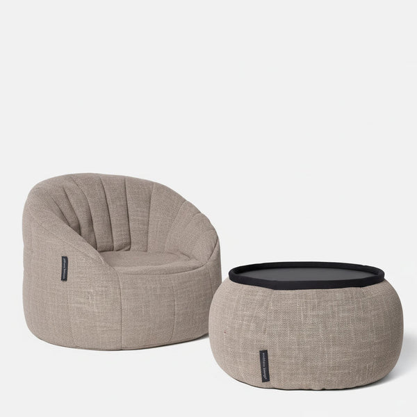 Beige, verhoiltu lounge-tuoli, jossa on pyöristetty selkänoja, istuu vierekkäin sopivan pyöreän ottomaanin kanssa, jonka päällä on musta tarjotin - tämä Ambient Lounge Cove -pakettiin kuuluva design-sittegruppe tuo mukavuutta møbler-tyyliin mihin tahansa ympäristöön valkoista taustaa vasten.
