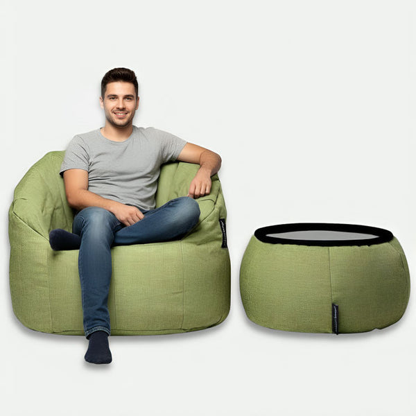 Hymyilevä mies, jolla on harmaa t-paita ja siniset farkut, istuu vihreällä Ambient Lounge®-papusäkkituolilla ja vihreällä ottomaanilla vaalealla taustalla.